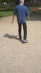 Back view of a person walking away on a gravel park path, wearing a blue shirt and dark pants with sneakers; benches and greenery in the background.